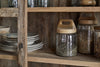 The Ibo Reclaimed Wood & Glass Cabinet - Natural - Wide by nkuku features antique brass handles and displays stacked ceramic bowls and jars of lentils or seeds, adding cozy, natural storage charm to your kitchen.