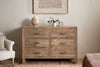 The nkuku Iya Reclaimed Wood Chest of Drawers - Natural - Large, featuring six drawers, stands against a light wall, topped with a textured vase, small plant, and clear box. An armchair and bed are partially visible on either side.