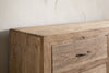 A close-up of the nkuku Iya Reclaimed Wood Chest of Drawers in natural—large size—features light reclaimed wood with visible grain and a single metal handle, set against a textured white wall for a rustic look.