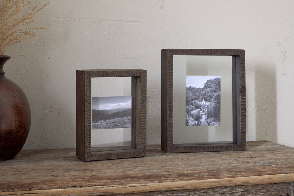 Two nkuku Indu Standing Mango Wood Frames in Dark Ash, each displaying black and white landscape photos, rest on a rustic wooden surface against a light wall. On the left, a brown vase with dried grass is partially visible.