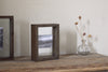 A black-and-white landscape photo is displayed in the nkuku Indu Standing Mango Wood Frame - Dark Ash, set on a wooden table beside a small glass vase with dried stems, all against a plain light-colored wall.