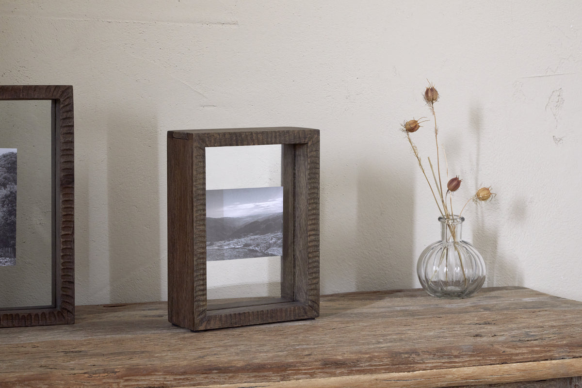 A black-and-white landscape photo is displayed in the nkuku Indu Standing Mango Wood Frame - Dark Ash, set on a wooden table beside a small glass vase with dried stems, all against a plain light-colored wall.