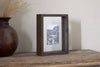 The nkuku Indu Standing Mango Wood Frame in Dark Ash displays a black-and-white photo on a rustic table next to a brown vase with dried grass, set against a light-colored textured wall.