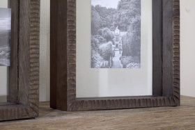 The Indu Standing Mango Wood Frame - Dark Ash by nkuku is shown on a wooden surface, displaying a black and white house photo. Another modern frame is partially visible on the left.