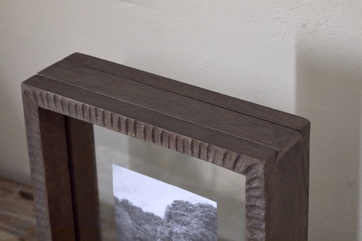 A close-up of the Indu Standing Mango Wood Frame - Dark Ash by nkuku, featuring a glass front and black-and-white photo of trees. Made from sustainable mango wood with a rustic finish, it rests against a light-colored wall.