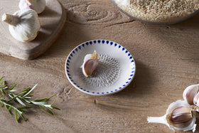 A wooden table features nkukus Indigo Drop Garlic Grater in Cream & Indigo with a clove of garlic, surrounded by garlic bulbs, rosemary sprig, a wooden board, and a glass jar of brown rice.