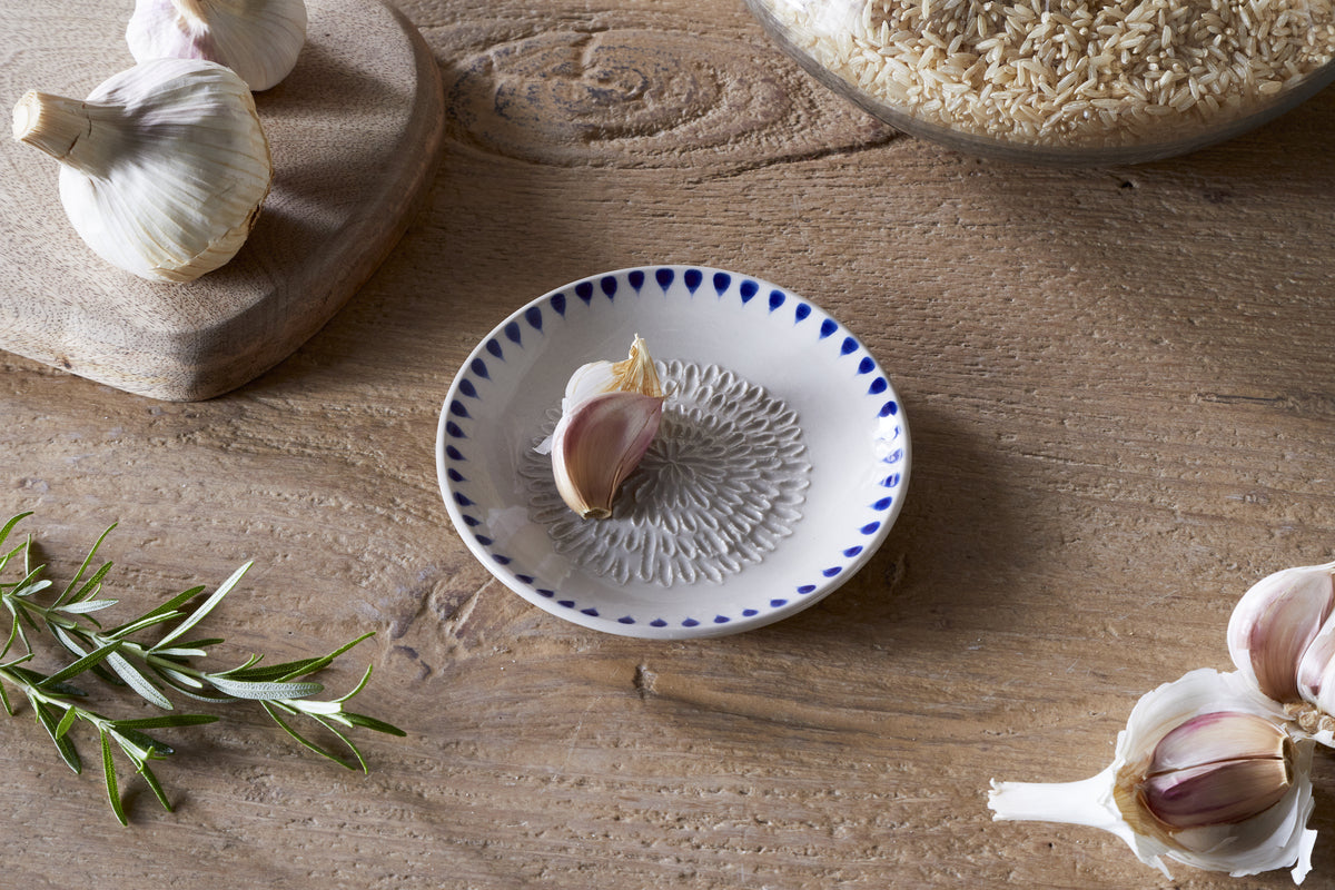 A wooden table features nkukus Indigo Drop Garlic Grater in Cream & Indigo with a clove of garlic, surrounded by garlic bulbs, rosemary sprig, a wooden board, and a glass jar of brown rice.