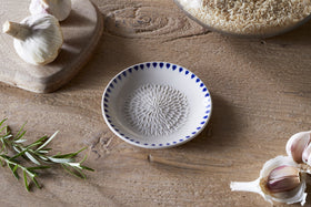 The nkuku Indigo Drop Garlic Grater - Cream & Indigo, a small white ceramic dish with a textured center and hand-painted indigo pattern, rests on a wooden table beside garlic, rice in a glass jar, a cutting board, and rosemary.