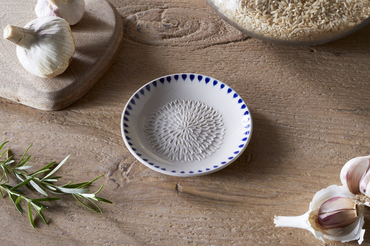 The nkuku Indigo Drop Garlic Grater - Cream & Indigo, a small white ceramic dish with a textured center and hand-painted indigo pattern, rests on a wooden table beside garlic, rice in a glass jar, a cutting board, and rosemary.