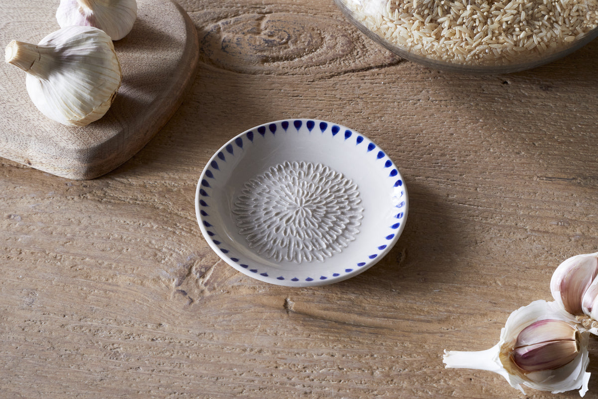 The nkuku Indigo Drop Garlic Grater - Cream & Indigo, with its hand-painted indigo design, rests on a wooden table among garlic bulbs, a cutting board, and a glass jar of rice.