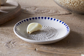 The nkuku Indigo Drop Garlic Grater in cream and indigo, featuring a textured center and hand-painted design, sits on a small white plate—perfect for grating garlic and adding style to your kitchen.