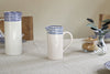 A small nkuku Indigo Drop Jug in cream and indigo sits on a linen tablecloth, surrounded by matching contemporary serveware, a sugar jar, and a glass vase filled with green leaves.