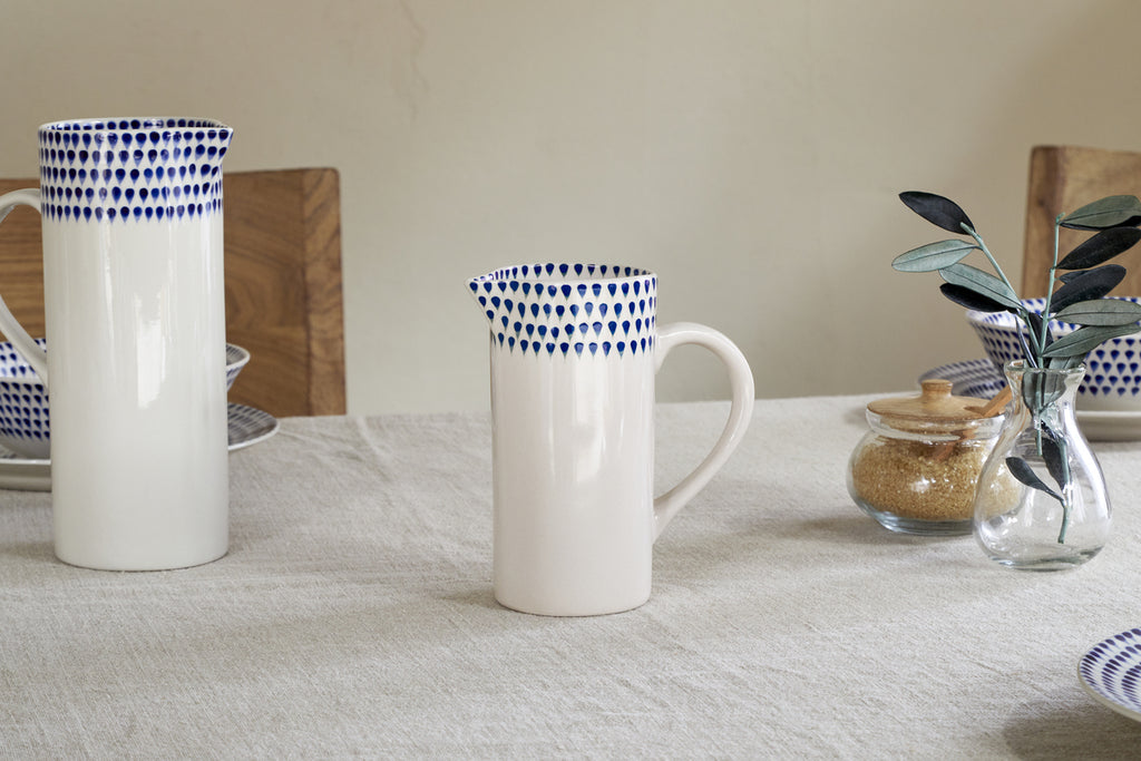A small nkuku Indigo Drop Jug in cream and indigo sits on a linen tablecloth, surrounded by matching contemporary serveware, a sugar jar, and a glass vase filled with green leaves.