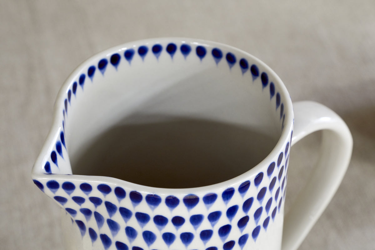 The nkuku Indigo Drop Jug - Cream & Indigo - Small features blue teardrop patterns along the rim. This white ceramic jug with a spout is ideal for contemporary serveware, displayed here on a neutral surface.