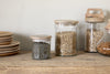 The Igoko Storage Jar - Clear by nkuku, filled with lentils, beans, and pasta, is neatly placed beside plates on a wooden countertop, creating a stylish kitchen organizer against a light-colored wall.