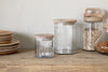 The Igoko Storage Jar - Clear by nkuku, with a mango wood lid, sits on a wooden surface beside stacked plates and jars of dried beans and pasta, creating a charming kitchen organizer against a light-colored wall.