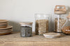 Nkuku’s Igoko Storage Jars—clear glass with mango wood lids—are filled with dry goods and arranged on a wooden countertop beside stacked wooden plates, set against a plain light wall for a tidy kitchen organizer look.