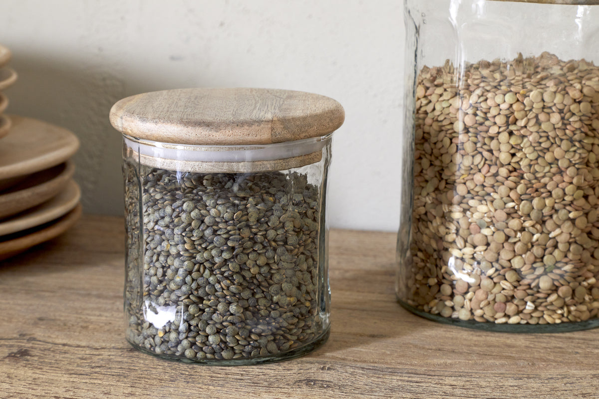 Two Igoko Storage Jars - Clear by nkuku, each with mango wood lids and filled with green and brown lentils, rest on a wooden surface beside stacked plates—an elegant way to organize your pantry.