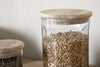 Two nkuku Igoko Storage Jars with mango wood lids sit on a wooden surface. The clear jar in front is filled with dried lentils, while the second holds another dried food. Perfect as a stylish kitchen organizer.