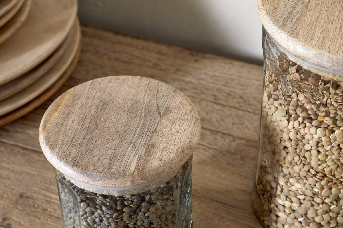 Two nkuku Igoko Storage Jars - Clear, with mango wood lids and filled with lentils, sit on a wooden surface beside stacked wooden plates, creating an elegant kitchen organizer.