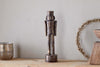 The nkuku Imay Nutcracker in antique brass stands on a wooden surface, accompanied by a silver candle holder, wreath, and bowl—creating an inviting seasonal display against a light gray wall.