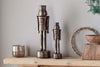 The Imay Nutcracker - Antique Brass by nkuku, in tall and short sizes, stands on a wooden shelf with a metallic candle holder, wooden bowl, and green foliage for a rustic, inviting seasonal display.