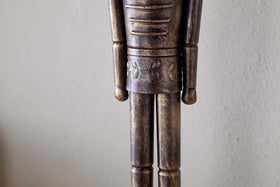 A close-up of the Imay Nutcracker - Antique Brass by nkuku shows its textured dark brown and gold finish, stylized cylindrical arms, and decorative waist carvings—perfect for festive or seasonal displays.