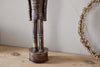 The Imay Nutcracker by nkuku, featuring an antique brass finish and elongated body, stands on a light wooden surface beside a dried floral wreath against a textured wall, creating a charming seasonal display.