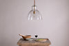 The Inayah Recycled Glass Pendant - Clear by nkuku hangs above a rustic wooden table set with a wooden bowl and pestle, gray bowl, glass of red wine, wooden board, knife, and sliced green vegetables against a plain light wall.