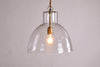 The Inayah Recycled Glass Pendant - Clear by nkuku features a dome-shaped, textured glass shade hanging from a gold fixture, illuminating the plain off-white wall with a warm, inviting glow.