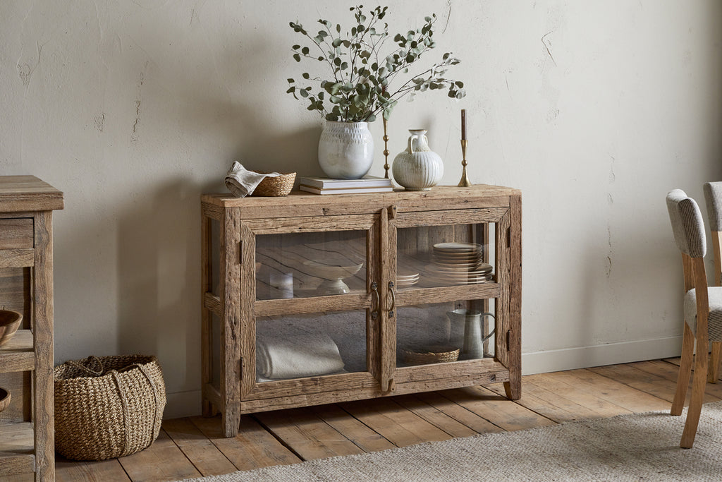 The nkuku Ibo Reclaimed Wood & Glass Sideboard - Natural showcases dishes behind glass doors with antique brass handles. Vases, candlesticks, and books adorn the top, while a woven basket rests on the wooden floor beside it.