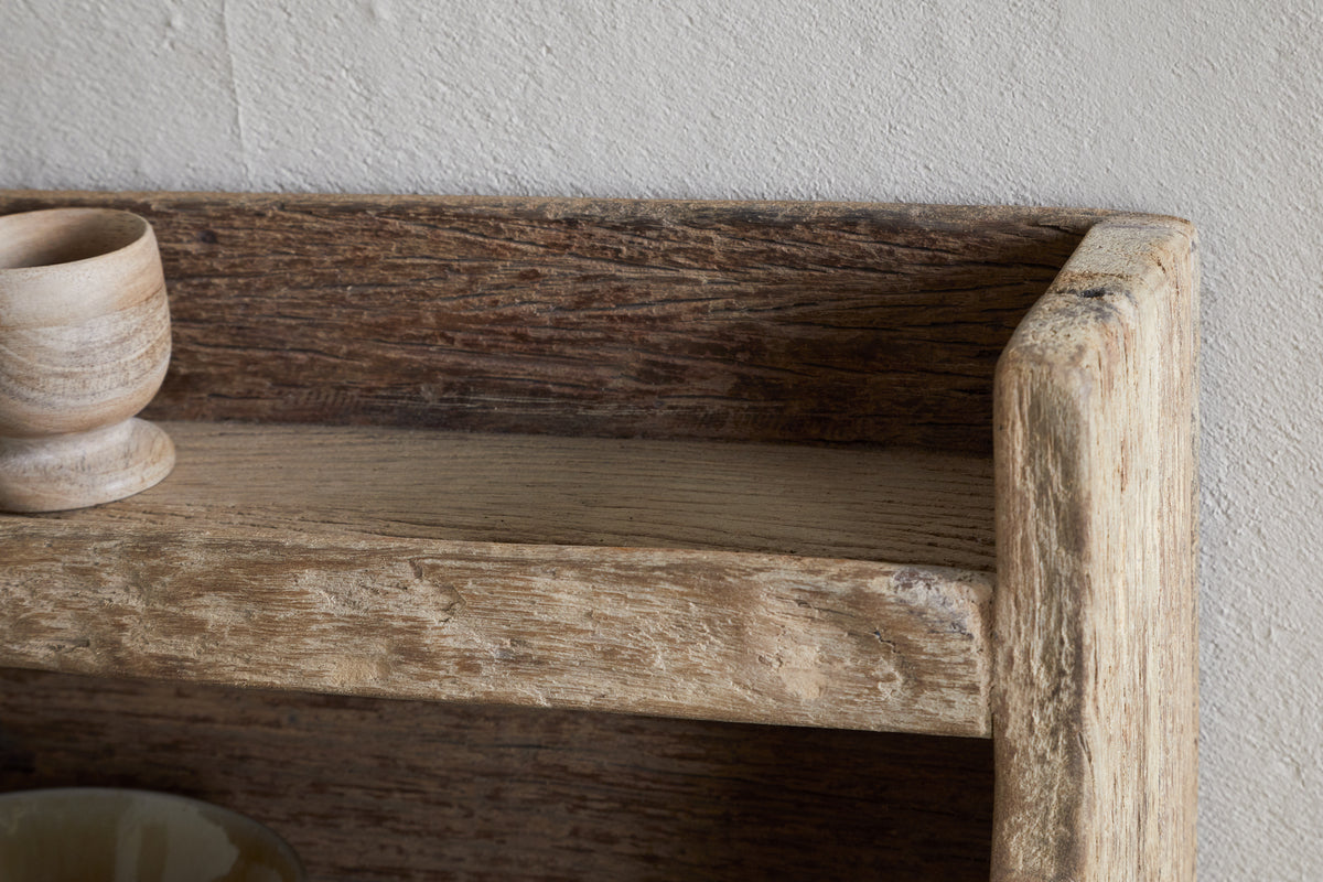Close-up of the nkuku Ibo Reclaimed Wood Storage Shelf - Natural, highlighting its weathered wood texture, with a small wooden cup on the left. This rustic wall-hung shelf adds charming kitchen storage against a light textured wall.
