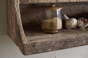 The Ibo Reclaimed Wood Storage Shelf by nkuku holds a glass jar with a wooden lid filled with brown sugar, plus garlic bulbs and cloves—ideal for stylish kitchen storage in a rustic setting.
