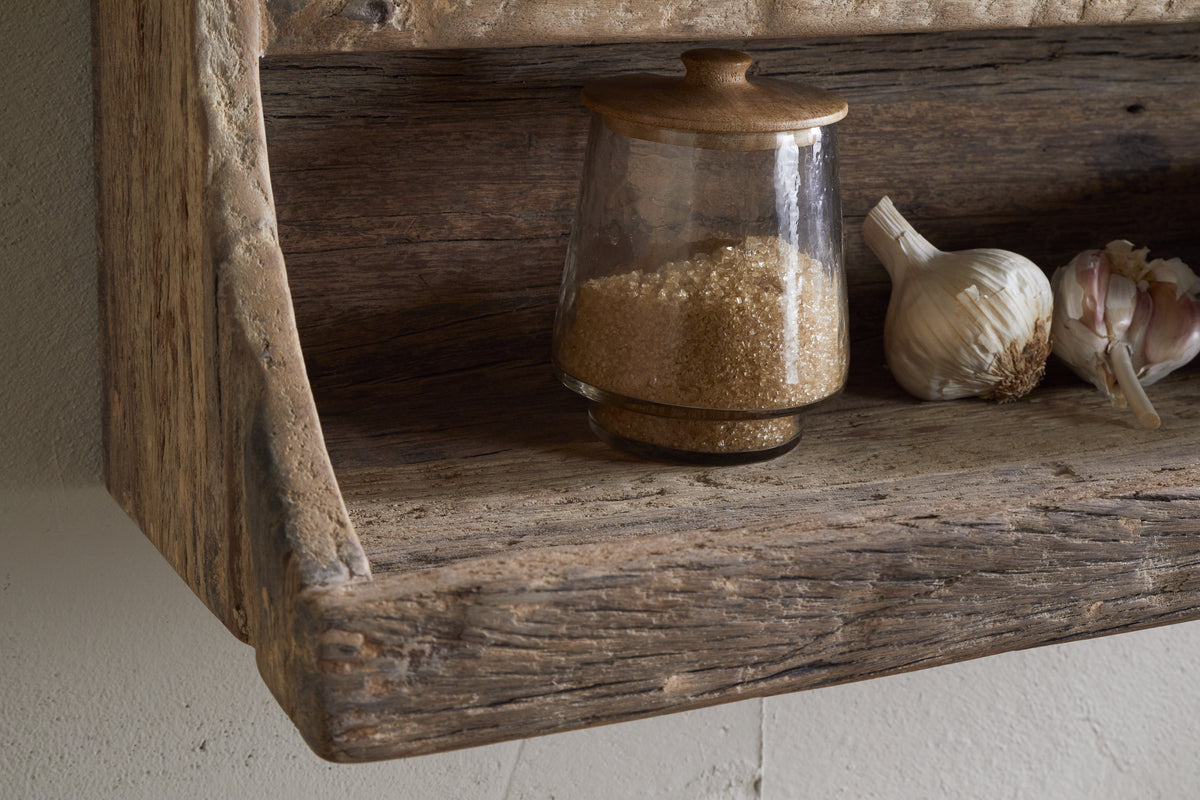The Ibo Reclaimed Wood Storage Shelf by nkuku holds a glass jar with a wooden lid filled with brown sugar, plus garlic bulbs and cloves—ideal for stylish kitchen storage in a rustic setting.