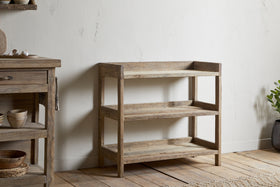 The nkuku Ibo Reclaimed Wood Pantry Standing Shelves in Natural, featuring a rustic three-tier open design, stands against a white textured wall on a wooden floor. A matching cabinet with bowls and a small potted plant complete the scene.