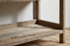 A close-up of the lower shelf and legs of the nkuku Ibo Reclaimed Wood Pantry Standing Shelves in Natural, resting on a light wooden floor against a white wall. The wood features a weathered, rustic finish.