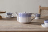 The nkuku Indigo Drop 4 Person Teapot in Cream & Indigo, adorned with hand-painted blue dots, sits on a wooden table with matching teacups and saucers—an ideal gift for tea lovers. A woven chair appears in the background.