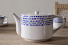 The nkuku Indigo Drop 4 Person Teapot in cream & indigo, featuring a blue dotted pattern, rests on a wooden table with matching cups and a woven-back chair in the background—an ideal gift for tea lovers.