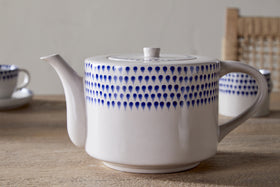 The nkuku Indigo Drop 4 Person Teapot in cream & indigo, featuring a blue dotted pattern, rests on a wooden table with matching cups and a woven-back chair in the background—an ideal gift for tea lovers.
