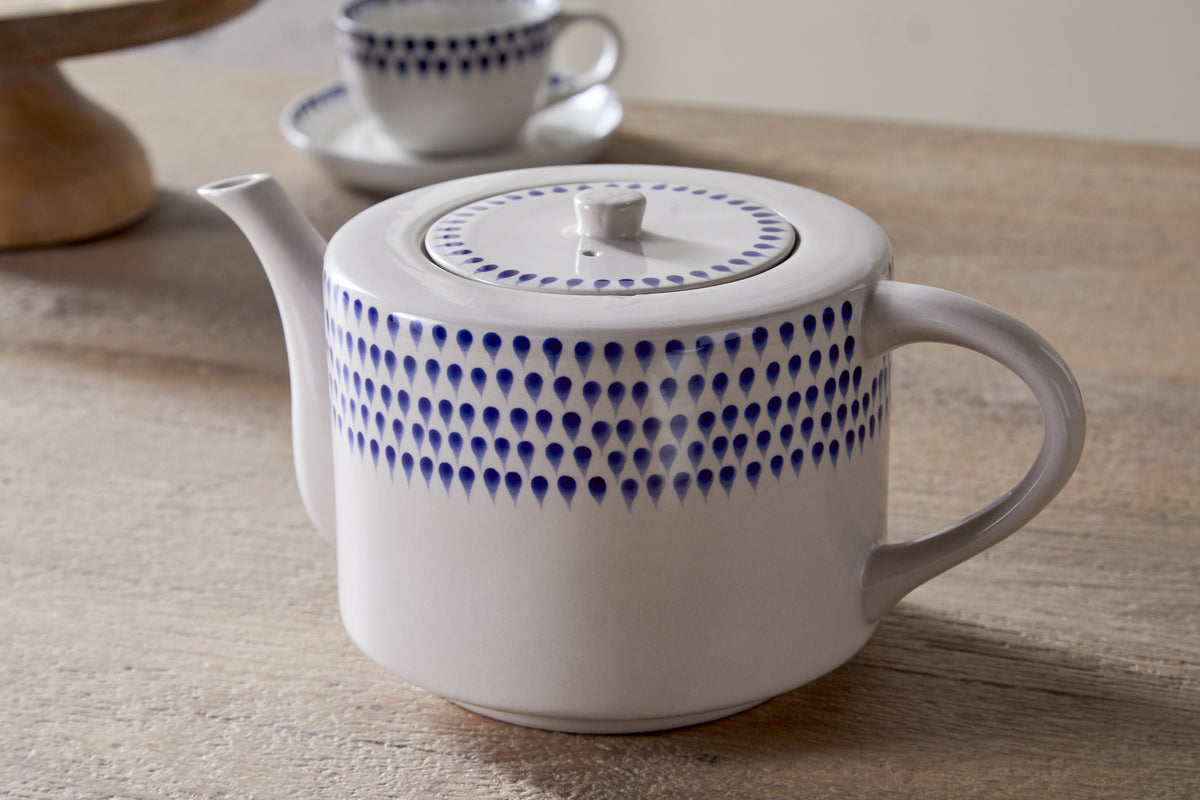 The nkuku Indigo Drop 4 Person Teapot in cream & indigo, featuring hand-painted blue teardrop dots, sits on a wooden surface. A matching cup and saucer are blurred in the background—perfect for tea lovers.
