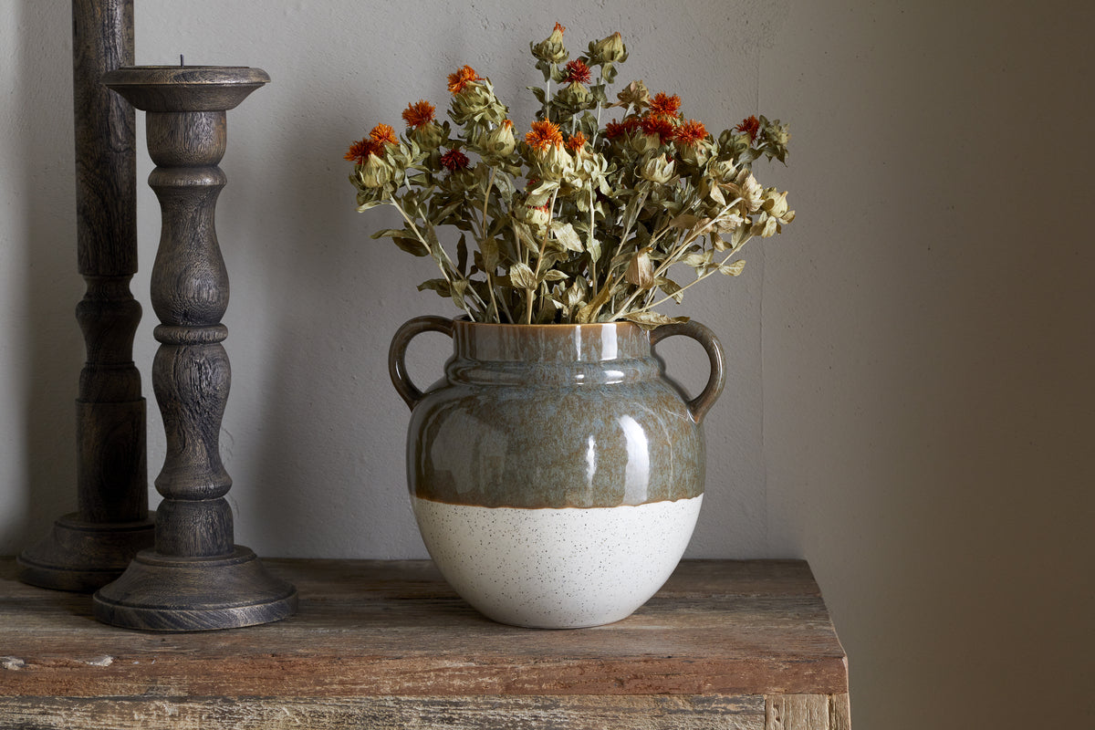 The nkuku Inha Reactive Glaze Vase in Dark Olive & Natural, featuring two handles, displays dried orange and beige flowers. It sits on a weathered wood surface beside dark candle holders against a plain light wall.
