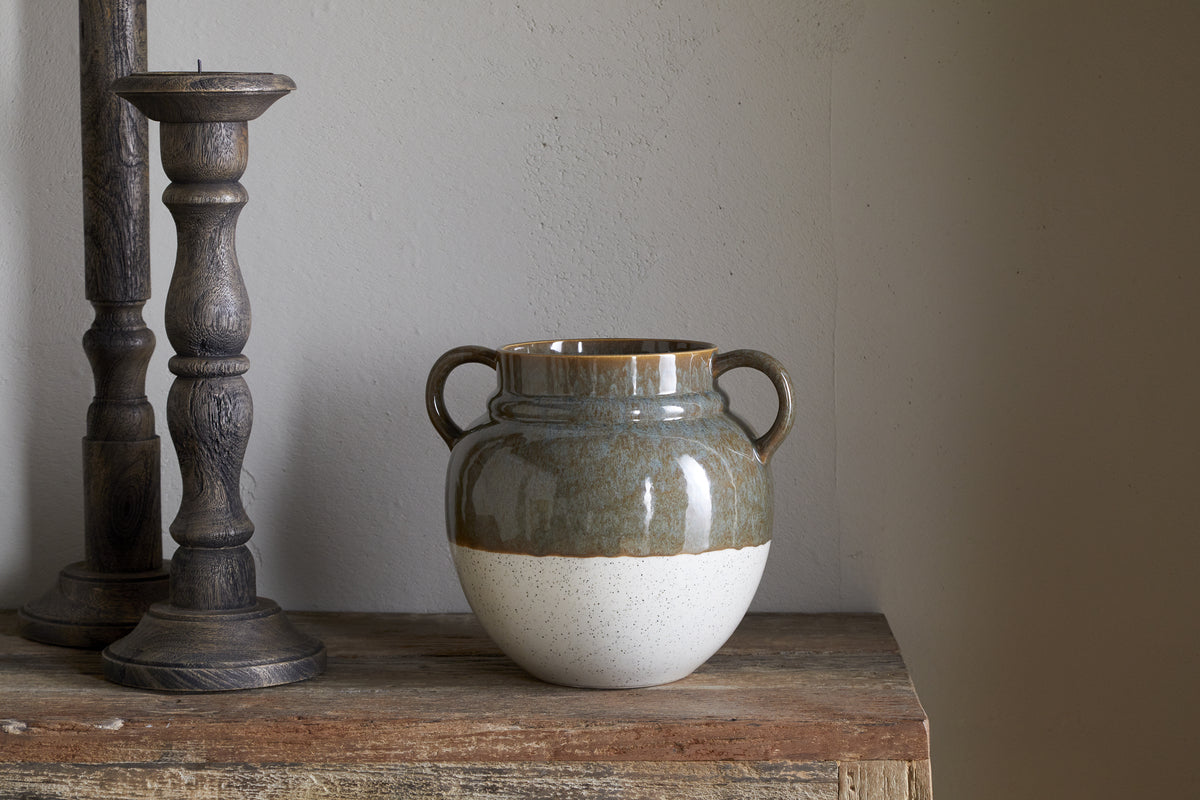 The nkuku Inha Reactive Glaze Vase in Dark Olive & Natural, with two handles and a brown-green glossy top over a speckled off-white base, sits on rustic wood, flanked by two carved dark wooden candle holders against a light gray wall.