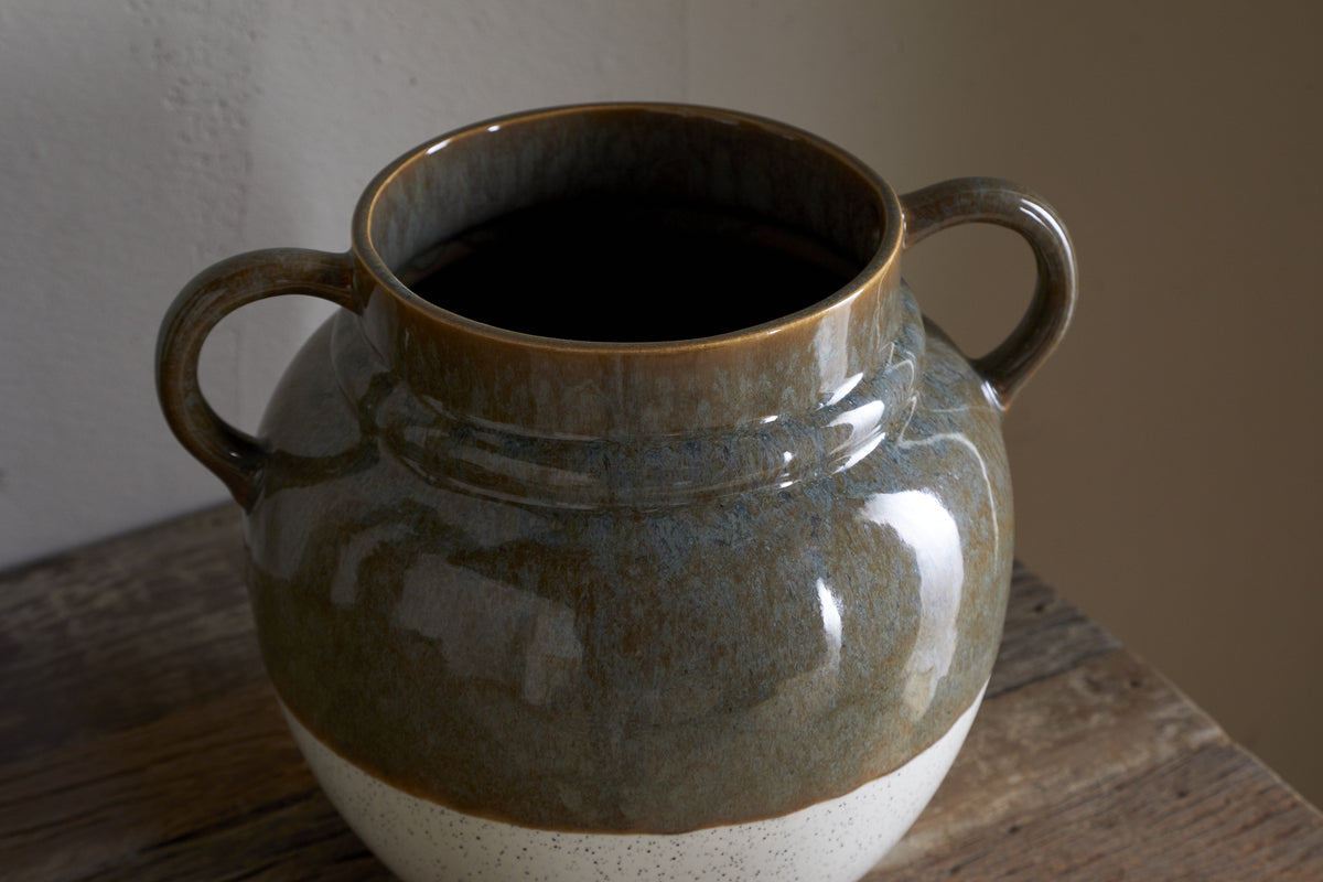 The nkuku Inha Reactive Glaze Vase in Dark Olive & Natural features two handles and sits on a rustic wooden surface, showcasing a glossy dark olive upper half, a matte speckled white base, and smooth texture highlighted by light and shadow.