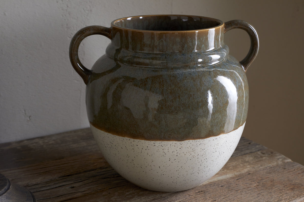 The Inha Reactive Glaze Vase by nkuku sits on a wooden surface, featuring two handles. Its top half is glossy dark olive, while the speckled matte bottom is natural off-white, with reflections and shadows enhancing its smooth texture.