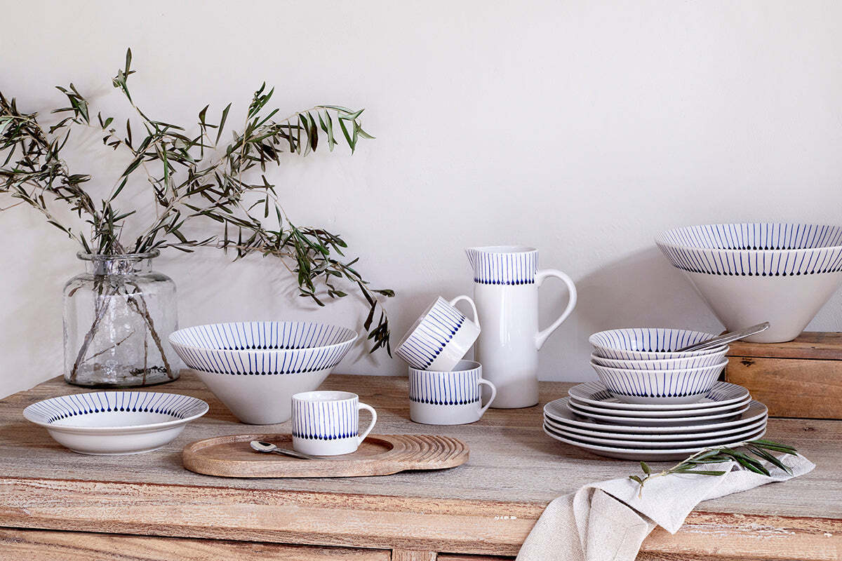 A rustic wooden table displays nkuku's handmade white clay dishes with indigo blue lines—bowls, plates, cups, and the Iba Jug - Indigo. Olive branches in a glass jar and a beige napkin by stacked plates highlight artisanal craftsmanship.
