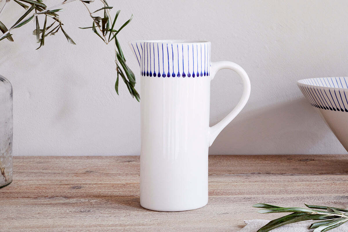 The Iba Jug - Indigo by nkuku, a tall white ceramic jug with vertical blue lines and dotted details near the rim, rests on a wooden surface beside olive branches and a matching bowl in a minimal, natural setting.
