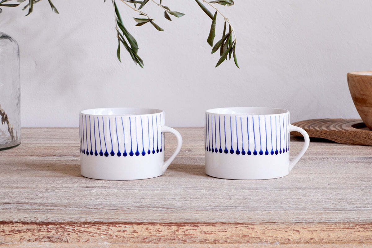 A set of two large nkuku Iba Mugs in indigo, featuring blue vertical stripes and teardrop bottoms, rest on a light wood table with green olive branches in the background.