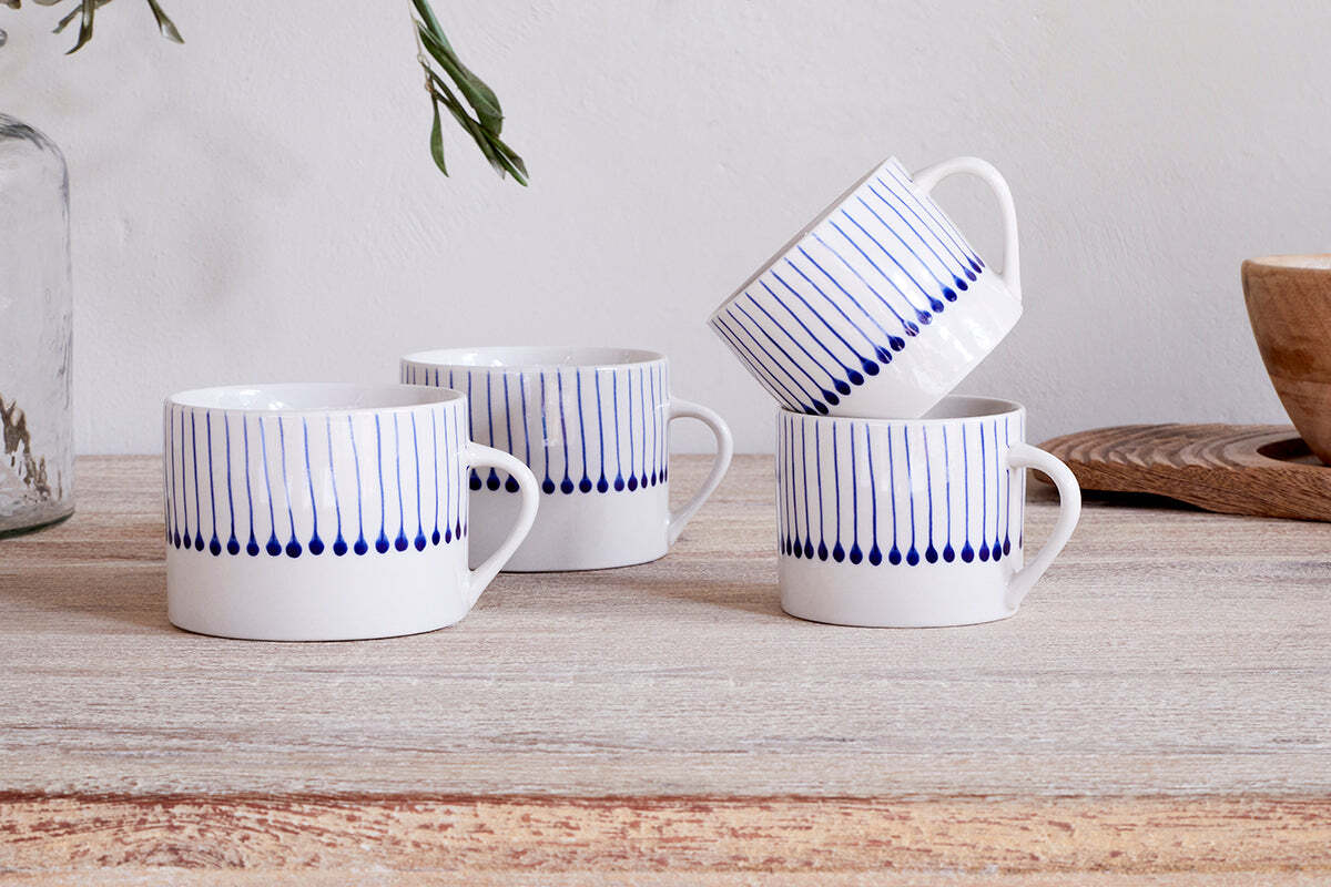 Two sets of nkuku Iba Mug - Indigo - Large (Set of 2) feature white mugs with vertical blue lines and teardrop designs, arranged on a light wooden table; three stand upright, one rests at an angle, with glass bottle and bowl in the background.