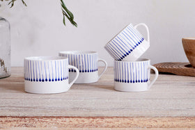Two sets of nkuku’s Iba Mug - Indigo - Small (Set of 2) with hand-painted blue raindrop stripes rest on a rustic wooden surface, beside a wooden bowl and glass vase with greenery in the background.
