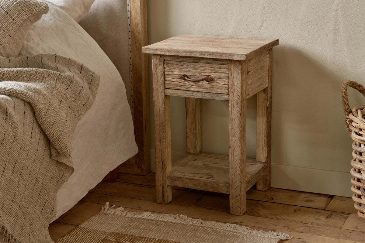 The nkuku Ibo Reclaimed Wood Bedside Table - Natural, featuring a single drawer with a metal handle and a lower shelf, sits on a wooden floor beside neutral bedding, a woven basket, and a textured rug for a cozy look.